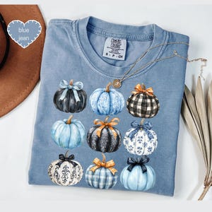 May include: A blue Comfort Colors t-shirt featuring nine watercolor pumpkins with bows. The pumpkins are in various colors and patterns, including black, blue, and plaid. A gold necklace is draped across the shirt.