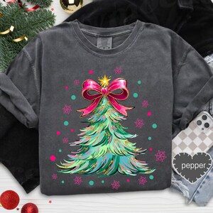 May include: Dark gray sweatshirt featuring a Christmas tree design. The tree is green and teal with a pink bow and gold accents. Pink and teal dots and snowflakes surround the tree. A gold star sits atop the tree.