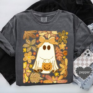 May include: A dark gray sweatshirt featuring a whimsical ghost design holding a jack-o'-lantern. The ghost is surrounded by a floral arrangement in shades of orange, yellow, and brown. A large bow adorns the ghost's head, adding a touch of charm.