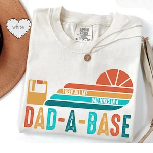 May include: White t-shirt with the text "I KEEP ALL MY DAD JOKES IN A DAD-A-BASE" in orange, teal, and yellow. The shirt features a retro design with a floppy disk and a sliced orange graphic.