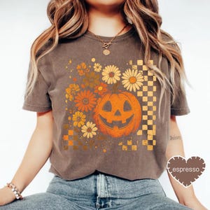 May include: Brown t-shirt featuring a smiling jack-o'-lantern graphic with flowers and a checkered pattern. The design incorporates orange, yellow, and brown tones, evoking a vintage Halloween aesthetic. The word "espresso" is printed on a heart-shaped tag.