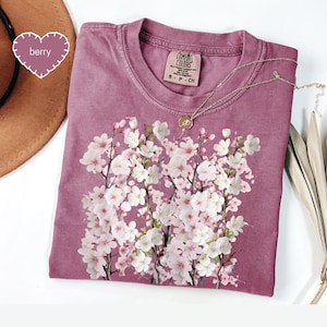 May include: Berry-colored t-shirt with a floral design of white and pink blossoms. The shirt has a crew neck and short sleeves. A silver necklace with a small pendant rests on the shirt. The tag reads "Comfort Colors".
