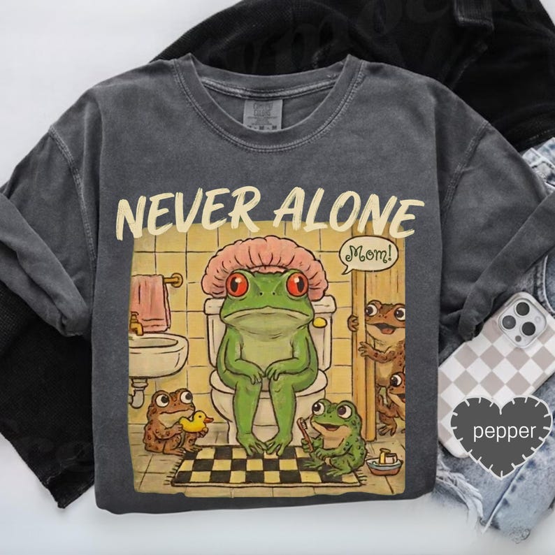 a gray t-shirt with a cartoon illustration of a frog sitting on a toilet, surrounded by other cartoon frogs, with a speech bubble saying "Mom!".