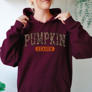 A person wearing a maroon hoodie with the word "PUMPKIN" and "SEASON" printed on it.
