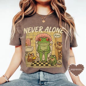 A woman wearing a t-shirt with a cartoon image of a frog sitting on a toilet and a speech bubble saying "Mom!".