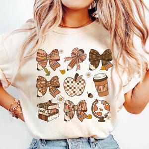 May include: Cream-colored t-shirt featuring a collection of fall-themed graphics. The design includes bows, pencils, an apple, a coffee cup, books, and a globe. The graphics are in shades of brown, orange, and cream, with accents of white and green.