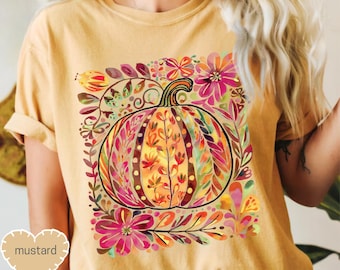 May include: Mustard-colored t-shirt featuring a colorful pumpkin design. The pumpkin is surrounded by floral elements in shades of pink, orange, and green. The word "mustard" is printed on a heart-shaped tag.