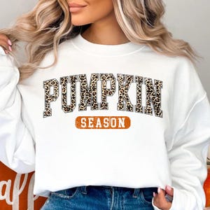 May include: White sweatshirt with the text "PUMPKIN" in a leopard print design above the word "SEASON" in an orange rectangle. The sweatshirt is a fall-themed apparel item.