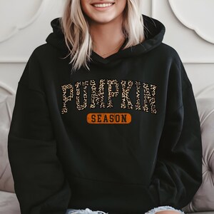 A woman wearing a black hoodie with the word "PUMPKIN" printed on it, sitting on a couch.