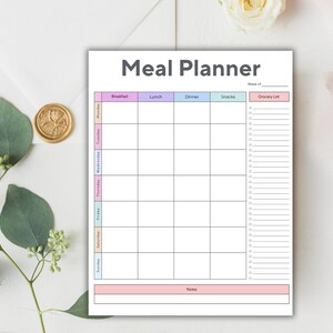 Op de afbeelding: Een witte maaltijdplanner met de titel "Meal Planner" in grijs. De planner heeft secties voor elke dag van de week, met kolommen voor ontbijt, lunch, diner, snacks en een boodschappenlijst. De planner is versierd met pastelkleuren.