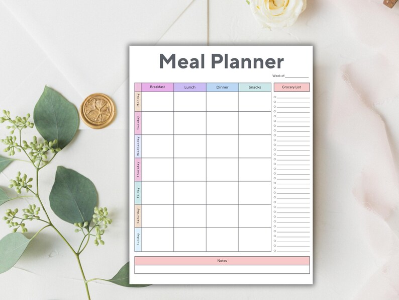 Op de afbeelding: Een witte maaltijdplanner met de titel "Meal Planner" in grijs. De planner heeft secties voor elke dag van de week, met kolommen voor ontbijt, lunch, diner, snacks en een boodschappenlijst. De planner is versierd met pastelkleuren.