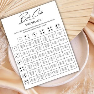 May include: A white printed sheet titled "Book Club Dice Breaker" with discussion questions. The sheet is on a white plate, with a beige background. The text includes instructions to pass the dice around the circle and dive into a fresh discussion question about the book.