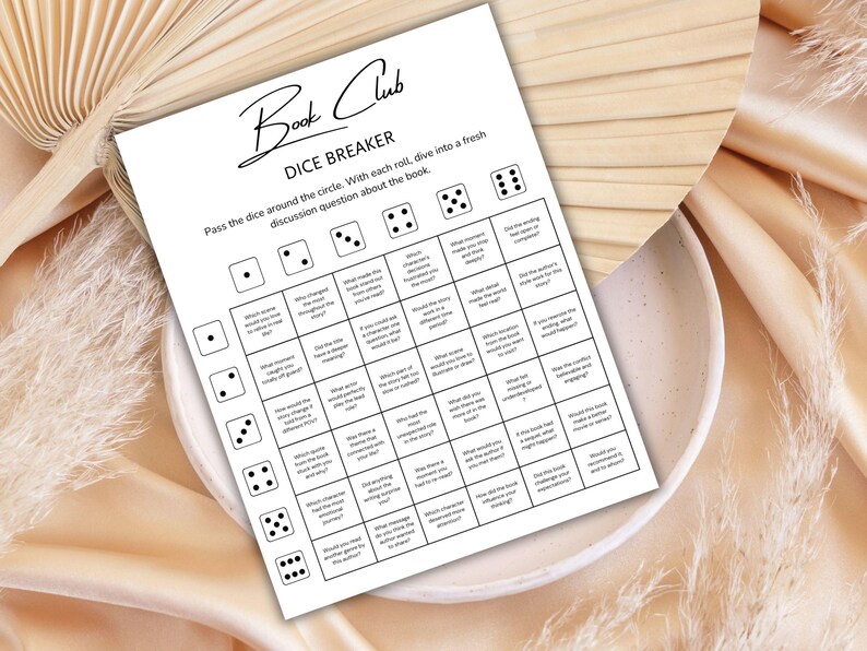 May include: A white printed sheet titled "Book Club Dice Breaker" with discussion questions. The sheet is on a white plate, with a beige background. The text includes instructions to pass the dice around the circle and dive into a fresh discussion question about the book.