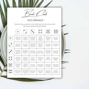 May include: A white Book Club Dice Breaker game sheet with discussion questions. The sheet has a grid with dice images and questions. The text "Book Club" is at the top in a script font. The sheet is on a plate with green leaves.