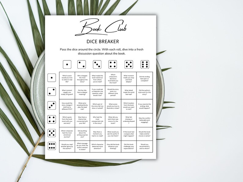 May include: A white Book Club Dice Breaker game sheet with discussion questions. The sheet has a grid with dice images and questions. The text "Book Club" is at the top in a script font. The sheet is on a plate with green leaves.