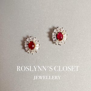 以下が含まれることがあります： 中央に赤い宝石、周囲に白と透明な石をあしらったスタッドピアス2つ。ピアスは明るいグレーの背景に配置されています。「ROSLYNN'S CLOSET JEWELLERY」の文字が下にあります。