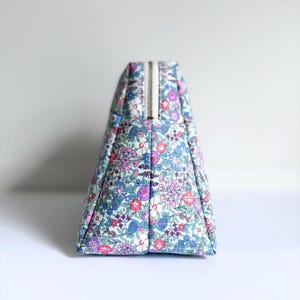 Könnte beinhalten: Ein dreieckiges Täschchen mit einem dichten Blumenmuster aus kleinen rosa, violetten, blauen und weißen Blüten auf hellblauem Grund. Das Täschchen verfügt über einen metallischen Reißverschluss an der Oberkante und einen strukturierten, flachen Boden.