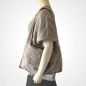 May include: A large, brown, fabric tote bag with a unique, textured design. The bag is made of multiple layers of fabric, creating a layered, patchwork effect. The bag is worn over the shoulder of a person wearing a brown linen top.