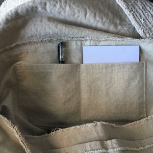 May include: A beige canvas tote bag with a zippered top and two interior pockets. A black pen and a white piece of paper are in the pockets.