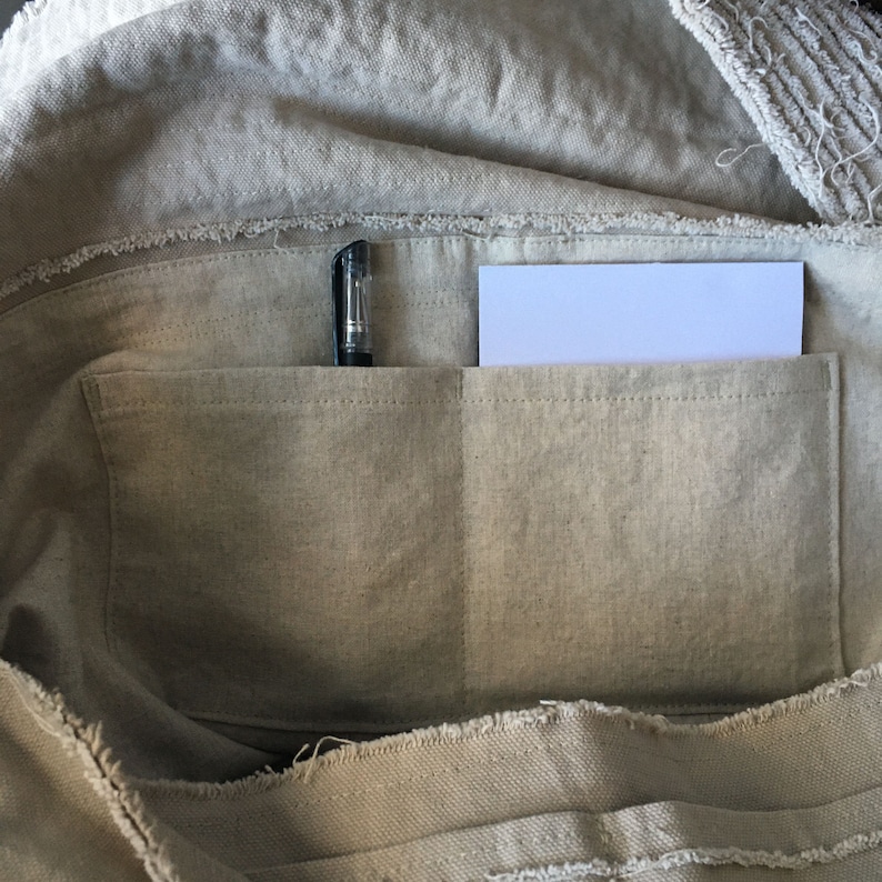 May include: A beige canvas tote bag with a zippered top and two interior pockets. A black pen and a white piece of paper are in the pockets.