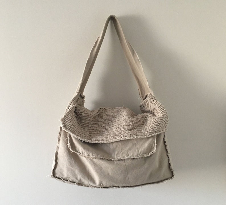 May include: A beige canvas tote bag with a frayed edge and a single strap. The bag has a folded over top with a textured fabric.