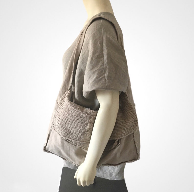 May include: A large, brown, fabric tote bag with a unique, textured design. The bag is made of multiple layers of fabric, creating a layered, patchwork effect. The bag is worn over the shoulder of a person wearing a brown linen top.