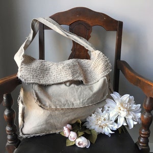 May include: A beige canvas tote bag with a frayed edge and a textured fabric flap. The bag is sitting on a wooden chair with white flowers in front of it.