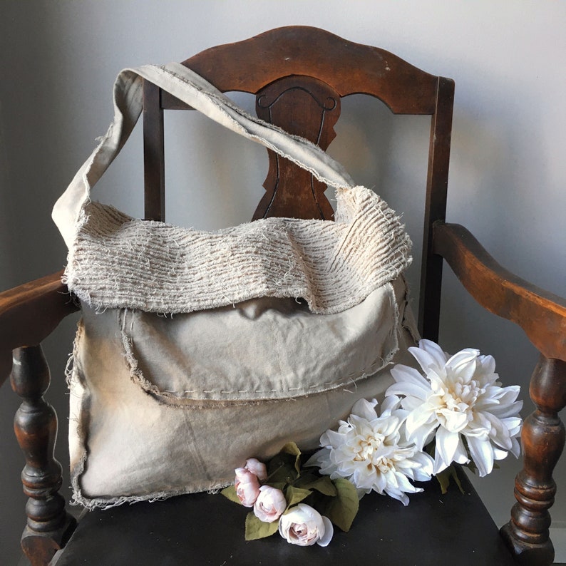 May include: A beige canvas tote bag with a frayed edge and a textured fabric flap. The bag is sitting on a wooden chair with white flowers in front of it.