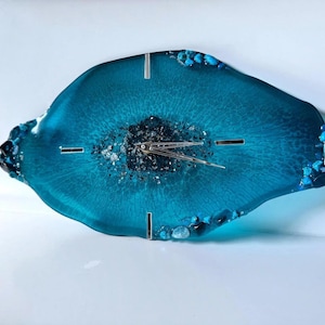 May include: An irregular, geode-inspired wall clock in translucent teal resin. The center features dark, textured mineral accents, while the edges are adorned with small, embedded blue and clear stones. It has silver clock hands and markers.