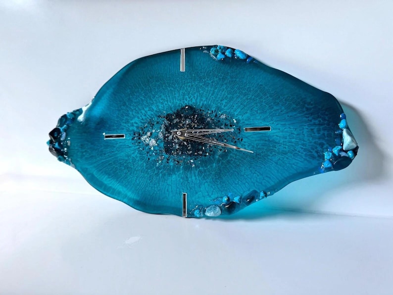 May include: An irregular, geode-inspired wall clock in translucent teal resin. The center features dark, textured mineral accents, while the edges are adorned with small, embedded blue and clear stones. It has silver clock hands and markers.