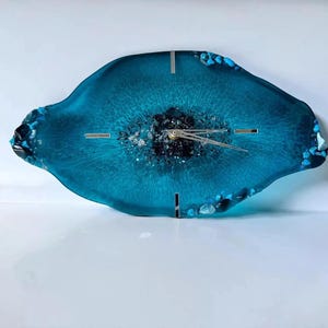 May include: An irregular, oval-shaped wall clock made of translucent teal resin. The center features dark, textured mineral-like accents, while the edges are embedded with small, bright blue stones. It has silver clock hands and silver markers.