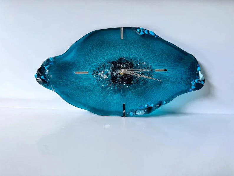 May include: An irregular, oval-shaped wall clock made of translucent teal resin. The center features dark, textured mineral-like accents, while the edges are embedded with small, bright blue stones. It has silver clock hands and silver markers.