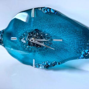 May include: An irregular, geode-inspired wall clock made of translucent teal resin. The center features dark, textured mineral accents surrounding silver clock hands. The edges are decorated with small, polished blue and clear stones.