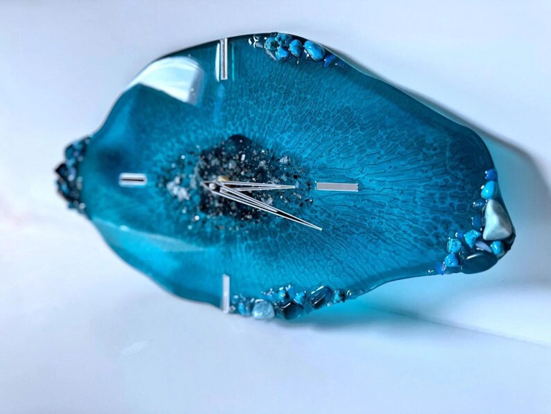 May include: An irregular, geode-inspired wall clock made of translucent teal resin. The center features dark, textured mineral accents surrounding silver clock hands. The edges are decorated with small, polished blue and clear stones.