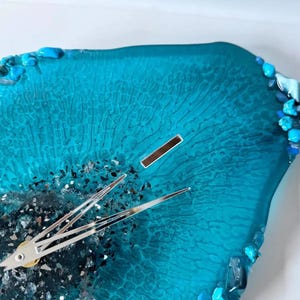 May include: A wall clock with a translucent teal resin face featuring a textured, web-like pattern. The center is accented with dark, crushed metallic fragments, and the edges are decorated with small, polished turquoise and dark blue stones.