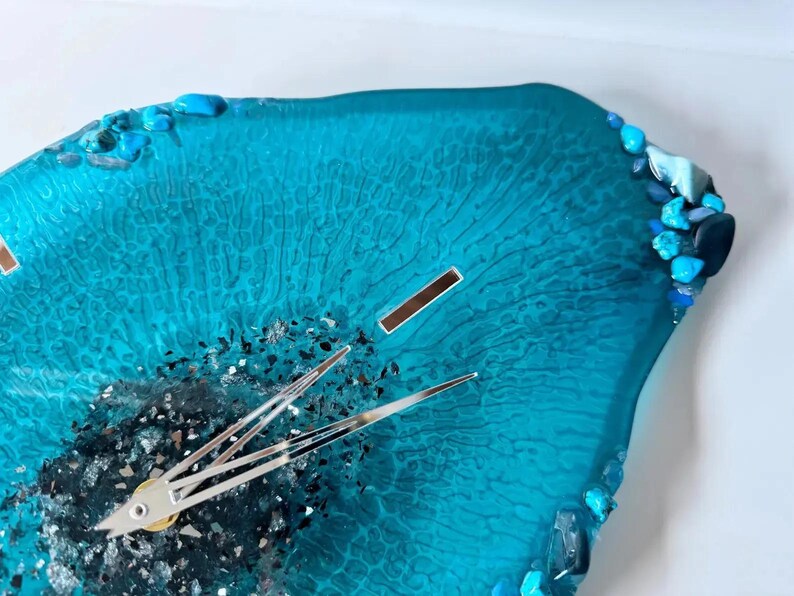 May include: A wall clock with a translucent teal resin face featuring a textured, web-like pattern. The center is accented with dark, crushed metallic fragments, and the edges are decorated with small, polished turquoise and dark blue stones.