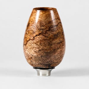 May include: A teardrop-shaped wooden vessel featuring a complex, swirling brown burl grain pattern with dark natural fissures. The object is mounted on a smooth, circular, metallic silver-toned base that provides a stable foundation.