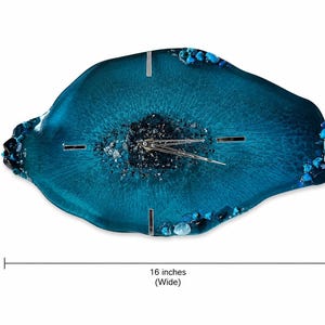 May include: An irregular, geode-inspired wall clock in shades of deep teal and blue resin. The center features dark crushed glass, surrounded by smooth resin and accented with blue stones along the edges. It measures 16 inches high by 16 inches wide.