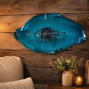 May include: A wall clock with an irregular, geode-inspired shape made of translucent teal resin. The center features dark, crushed mineral accents, while the edges are decorated with polished blue stones. It has silver hands and four silver markers.