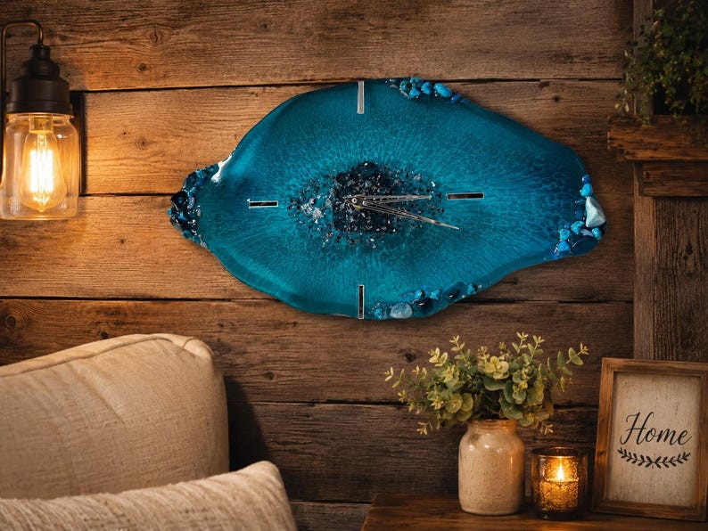 May include: A wall clock with an irregular, geode-inspired shape made of translucent teal resin. The center features dark, crushed mineral accents, while the edges are decorated with polished blue stones. It has silver hands and four silver markers.