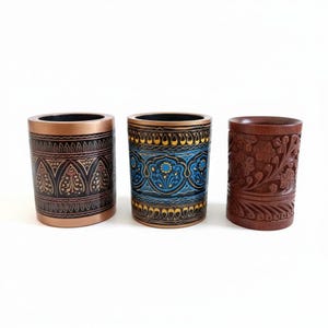 Può includere: Tre contenitori cilindrici decorativi. Il primo è color rame con motivi neri e oro. Il secondo è blu e oro con un motivo floreale. Il terzo è in legno intagliato con rilievo floreale.