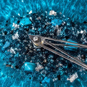 May include: A wall clock featuring a vibrant blue rippled glass base embedded with turquoise stones, silver leaf flakes, and dark glass shards. Two slender, polished silver metal clock hands are attached to a central silver-toned mounting post.
