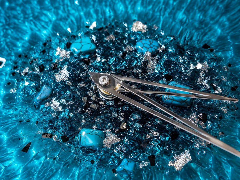 May include: A wall clock featuring a vibrant blue rippled glass base embedded with turquoise stones, silver leaf flakes, and dark glass shards. Two slender, polished silver metal clock hands are attached to a central silver-toned mounting post.