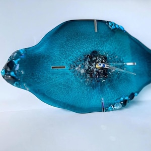May include: An oval-shaped wall clock made of translucent teal resin with a textured, dark center filled with metallic silver and black fragments. The clock features silver-toned hands and small, embedded blue and black stones along the edges.