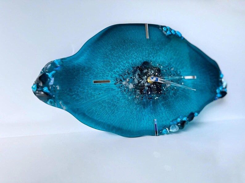 May include: An oval-shaped wall clock made of translucent teal resin with a textured, dark center filled with metallic silver and black fragments. The clock features silver-toned hands and small, embedded blue and black stones along the edges.