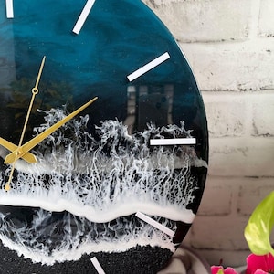 Könnte beinhalten: Eine runde Wanduhr mit einer glänzenden Harzoberfläche, die einen tiefblauen Ozean, weiße brechende Wellen und einen schwarzen Sandstrand darstellt. Die Uhr hat schmale weiße Stundenmarkierungen und dünne goldfarbene Metallzeiger für die Zeit.
