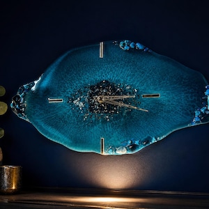 Può includere: Un orologio da parete di forma ovale con un corpo in resina blu profondo e un centro cristallino testurizzato. Il quadrante include lancette metalliche e indicatori delle ore dorati, accentuati da pietre blu incastonate lungo i bordi irregolari.