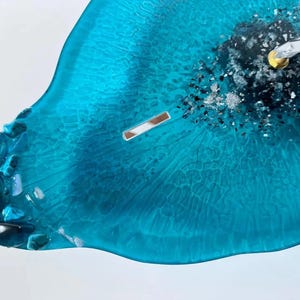 May include: A teal resin wall clock with a smooth, translucent finish and a textured, organic edge. The center features a cluster of dark gray and silver metallic flakes, with silver clock hands attached to a gold-toned central mechanism.