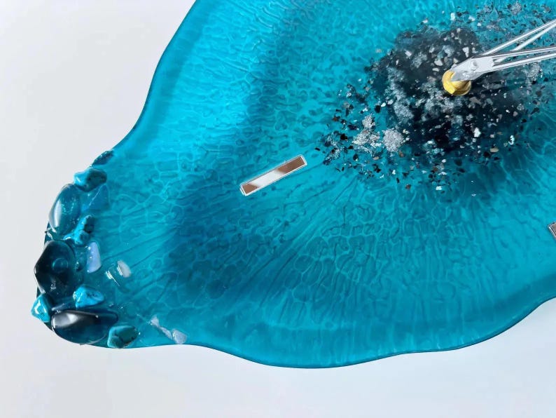 May include: A teal resin wall clock with a smooth, translucent finish and a textured, organic edge. The center features a cluster of dark gray and silver metallic flakes, with silver clock hands attached to a gold-toned central mechanism.