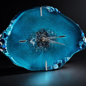 May include: An irregular, geode-inspired wall clock featuring a translucent, vibrant blue resin body. The center is filled with dark, shimmering crushed glass and metallic flakes. It includes silver-toned clock hands and three silver hour markers.